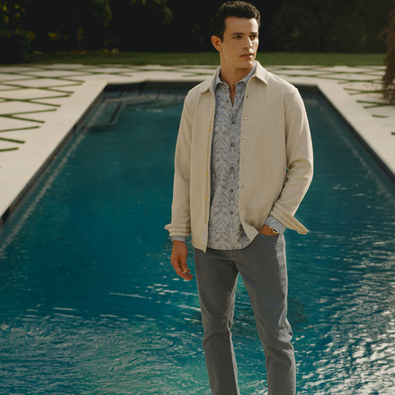 A man standing in front of a pool, wearing a light blue jacquard button up shirt with a cream shirt jacket layered over top