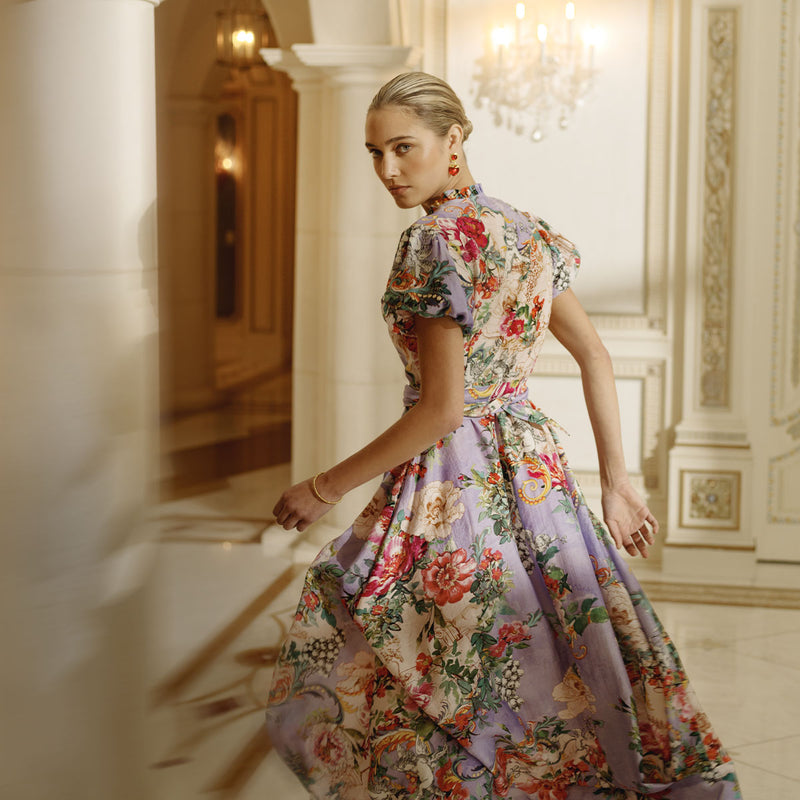 A woman in a purple floral print dress in a marble room looking over her shoulder